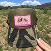 A hand holds up an olive green camouflage trucker hat with a pink 'FAITH' mountain patch, set against a sunny outdoor landscape of grassy fields and mountains under a blue sky.