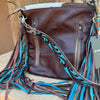 A brown leather shoulder bag with a wide, fringed strap featuring turquoise blue, brown, and black patterned strands, displayed on a stone surface next to a wooden background.