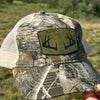 A camouflage trucker hat with a tan mesh back and a rectangular patch featuring black antlers on an olive background, held up by a hand against an outdoor grassy background.