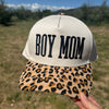 A hand holds a baseball cap outdoors in a grassy field. The cap has an off-white crown with black embroidered text reading 'BOY MOM' and a tan leopard-print brim with black and brown spots.