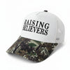 A white trucker-style cap with black text reading 'RAISING BELIEVERS' on the front panel and a camouflage-patterned brim, displayed on a stone surface outdoors.