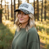 A white trucker-style cap with black embroidered text reading 'RAISING BELIEVERS' on the front, resting on a stone surface. The brim has a green and brown camouflage pattern.