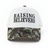 A white trucker-style cap with black text reading 'RAISING BELIEVERS' on the front, resting on a stone ledge outdoors. The brim has a green, black, and brown camouflage pattern.