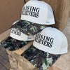 Three trucker-style caps with white mesh back panels and camouflage brims, each printed with the phrase 'RAISING BELIEVERS' in bold black text, displayed outdoors on a stone ledge.