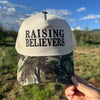 A hand holding a trucker hat outdoors with a grassy field and blue sky in the background. The hat has a cream-colored front panel with the text 'RAISING BELIEVERS' in black, and a camouflage-patterned brim.