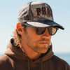 Man wearing a camouflage cap with 'PRAY' logo and sunglasses, standing against a clear sky.