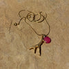 A gold necklace with a deer antler pendant and a round magenta pink stone, resting on a textured beige stone surface.