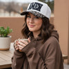 A woman wearing a brown hoodie and a white trucker hat with 'AMEN' in black letters and a leopard-print brim, holding a mug in an outdoor patio setting.