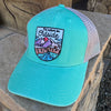 A turquoise trucker cap with a mesh back, featuring a colorful embroidered patch on the front that reads 'It rather be Outside' above a mountain and lake illustration. The cap is resting on a stone surface outdoors.