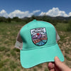 A teal green trucker hat with a colorful embroidered patch reading 'My favorite Outside' is held up by a hand against a blurred outdoor background of grass, mountains, and blue sky.