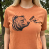 A close-up of a person wearing a heathered orange crewneck t-shirt with a black graphic of a bear's head and a pointing hand with the word 'BOOP!' printed on the chest, set against an outdoor background.