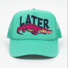 A bright turquoise trucker hat with a pink and yellow alligator graphic and the word 'LATER' in dark blue on the front, displayed on a stone surface outdoors.