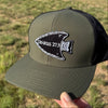 An olive green and black trucker hat with a black arrowhead patch embroidered with 'GENESIS 27:3' is held up by a hand against an outdoor grassy field background.