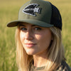 An olive green and black trucker hat with a black mesh back and a black arrowhead patch on the front, held by a hand against an outdoor grassy field background.