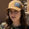 A tan baseball cap with a curved brim, featuring a black, white, and blue embroidered patch on the front that reads 'JESUS SAVES'. on a woman wearing camo