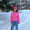 Person wearing a pink sweatshirt and hat standing in a snowy landscape with trees.