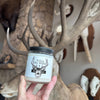 Hand holding a jar of candle in front of deer antlers and taxidermy heads.