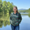 A woman wearing a green camouflage zip-up jacket with colorful floral patterns on the front, paired with blue jeans, standing in front of a brown wooden door and textured stucco wall.