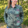 A woman wearing a dark green camouflage zip-up fleece jacket with pink and teal floral embroidery down the front, standing outdoors in front of a blurred green forest background.