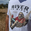A close-up of a person wearing a light gray heather t-shirt with a graphic print of a Bigfoot character lounging in a hammock under the text 'RIVER TO RIDGE'. The background is an outdoor dirt landscape.