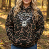 A person wearing a brown and green camouflage hoodie with a white graphic on the chest that reads 'Follow the WORD Not the herd' and shows blue jeans, standing in front of a wooden door and stucco wall.