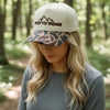 A trucker-style cap with an off-white crown and a brown camouflage-patterned brim. The front is embroidered with a mountain graphic and the words 'RIVER TO RIDGE' in brown.