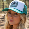 A teal and white trucker hat with a front patch reading 'FERAL BUT FORGIVEN' and showing a raccoon graphic, resting on a stone surface against a wooden wall.