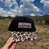 A hand holds a black baseball cap with a pink leopard-print brim and a front patch reading 'IT'S ABOUT TO GET WESTERN' against an outdoor desert landscape with blue sky and clouds.