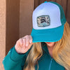 A woman wearing a teal and white trucker hat with a 'Feral but Forgiven' patch on the front, holding the brim down over her eyes. She is wearing a matching teal quarter-zip sweatshirt, standing in front of a stucco wall and wooden door.