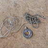 A bullet-shaped pendant necklace with a silver-toned casing and a gold-toned center, shown with two silver ball chains on a stone surface in front of a wooden wall.