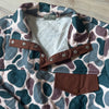 A close-up of a quilted pullover shirt by River to Ridge, featuring a brown, teal, and mauve camouflage pattern, a brown snap-button placket, and a matching brown chest pocket, laid flat on a light wood floor.