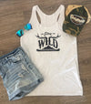 White tank top with 'Stay Wild' text, denim shorts, sunglasses, and cap on a wooden surface