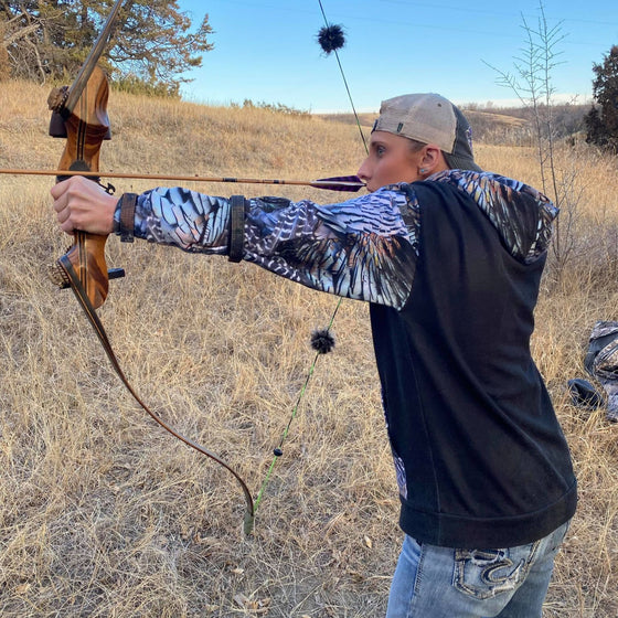 Woman shooting traditional archery with a bow and arrow and wearing a turkey feather hoodie from River to Ridge Clothing Brand