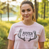 A woman wearing a light pink t-shirt with a black graphic that reads 'RIVER TO RIDGE' and features antler details, standing in a sunlit forest setting.