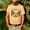 A bearded man wearing a beige graphic t-shirt with a forest and lake design, standing outdoors in front of green trees.