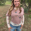 A woman standing outdoors in a grassy field with trees and mountains in the background, wearing a dusty rose cropped long-sleeve shirt with a duck hunting graphic print over a white long-sleeve undershirt and blue jeans.