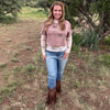 A woman stands outdoors in a grassy, tree-filled landscape wearing a dusty pink long-sleeve crop top with butterfly prints, blue jeans, and brown fringe boots.