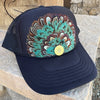 A black trucker hat with a colorful turquoise, brown, and white feather design on the front panel, resting on a stone surface outdoors.