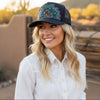 A smiling blonde woman wearing a navy blue trucker hat with turquoise and silver floral embroidery, paired with a white lace-trimmed button-up shirt, set against a warm desert landscape at sunset.