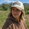 A woman outdoors wearing a beige trucker hat with 'RAISING BELIEVERS' printed on the front and a leopard-print brim, paired with a brown hoodie against a grassy field and blue sky.