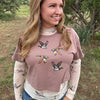 A woman wearing a dusty rose cropped t-shirt with a duck print over a cream long-sleeve shirt, standing outdoors in a grassy field with trees.