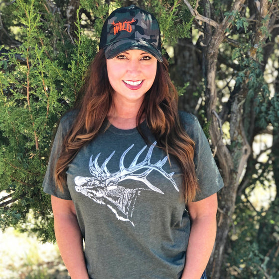 Woman with red hair wearing a green elk logo t shirt and a hunting hat in camo with the word wild on it in hunter orange. she is wearing jeans and standing in the forest