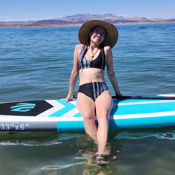 Woman on a paddleboard at lake mead in nevada, sitting in a high waist bikini that is black and grey with the USA flag on it, patriotic. She is smiling and wearing a straw hat