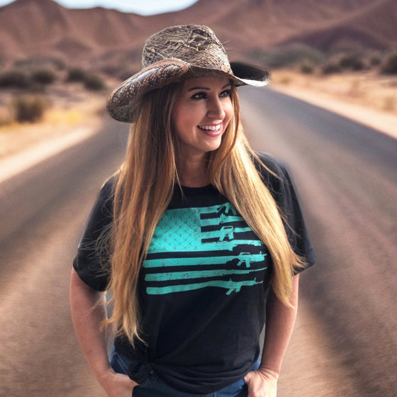 Woman on an open road in the desert wearing a cowboy hat and long red hair and wearing a teal gun flag 2A t shirt in black