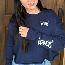  Woman with black hair wearing a cropped blue sweatshirt and jeans. The pullover has a WILD logo on with elk antlers for the letters