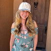 A smiling woman stands in front of a brown wooden front door wearing a distressed white baseball cap with 'AMEN' printed on it and a teal off-the-shoulder floral top.