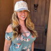 A smiling woman with long blonde hair stands in front of a brown wooden door, wearing a white baseball cap with 'AMEN' in gold letters and a dusty blue off-the-shoulder floral top.