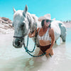Woman in a bikini and cowboy hat standing in water with a white horse.