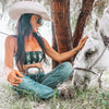 Woman in cowboy hat and sunglasses sitting next to a white horse in a natural setting.