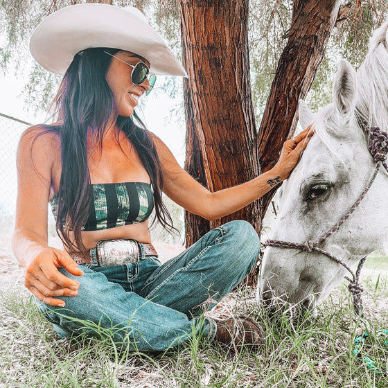 Woman in cowboy hat and sunglasses sitting next to a white horse in a natural setting.